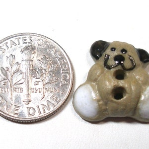 May include: A ceramic button shaped like a brown dog with black ears and a black nose. The dog has white feet and a black button for an eye.