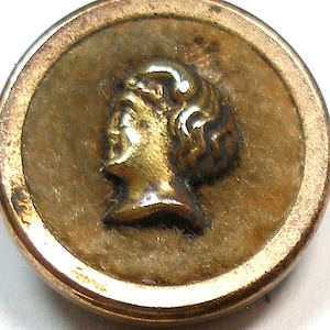May include: A vintage gold-toned button featuring a profile of a classical figure. The button has a circular design with a raised relief of a woman's face and hair. The button's edge has a gold border and a textured surface.
