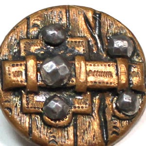 May include: A round, antique-style button with a bronze finish and silver-colored accents. The button features a textured surface with geometric designs and four silver-colored, faceted studs. The button is likely for sewing or crafts.