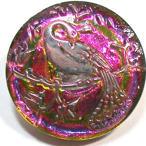 May include: A iridescent glass button with a raised design of a bird perched on a branch. The button has a pink, green, and blue color scheme.