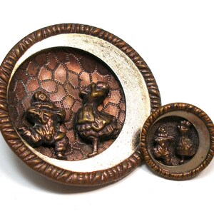 Peut inclure: Deux boutons anciens en bronze. Le plus grand bouton présente un relief détaillé de deux personnages sur un fond texturé de couleur cuivre, encadré par une bordure argentée et bronze. Un bouton plus petit reprend le motif.
