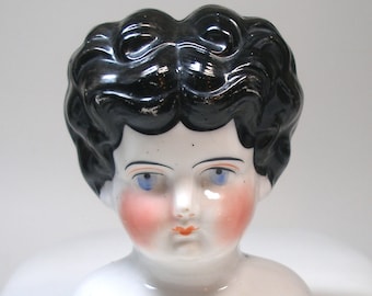May include: A porcelain doll head with black painted hair and blue eyes. The doll has painted rosy cheeks and a closed mouth.