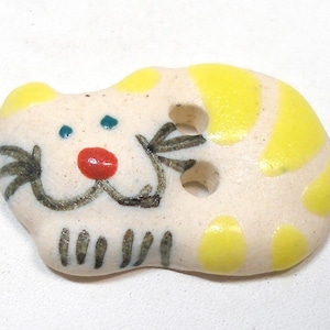 May include: A ceramic button in the shape of a cat. The button is off-white with yellow accents and a painted cat face featuring a red nose, blue eyes, and black whiskers. The button has two holes for sewing.