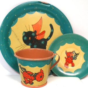 May include: A vintage children's tea set with a green and yellow color scheme. The set includes a cup, saucer, and plate. The cup and saucer feature a red and black dog with a red bow. The plate features a black cat with a red bow.