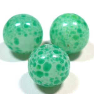 May include: Three green glass marbles with a speckled pattern. The marbles have a glossy finish and varying shades of green. The marbles are round and appear to be vintage or handmade.