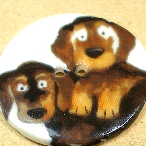 Puede incluir: Botón redondo y blanco con una ilustración pintada de dos perros. Los perros tienen pelaje marrón y negro con detalles blancos y ojos grandes y sorprendidos. El botón tiene dos agujeros para coser.