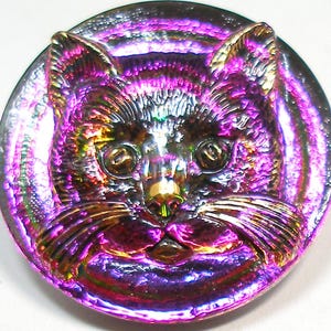 Puede incluir: Un botón de vidrio púrpura iridiscente con una imagen en relieve de la cara de un gato. El gato tiene pelaje negro y ojos dorados.