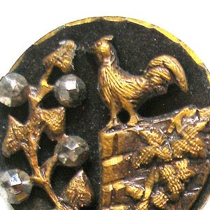 Puede incluir: Un botón vintage con un diseño detallado. El botón presenta un gallo dorado de pie sobre una pared de ladrillos, con una rama de cuentas claras y facetadas y hojas doradas. El fondo es negro.