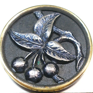 May include: A silver-toned metal button with a raised design of a cherry branch with three cherries and leaves. The button is set against a black background and has a gold-toned rim.