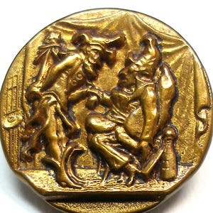 Op de afbeelding: Een vintage, ronde, messingkleurige knoop met een gedetailleerde reliëfscène. De knoop toont twee figuren in historische kleding, mogelijk een theatrale of historische scène. De figuren staan tegen een achtergrond met textuur, met ingewikkelde details.