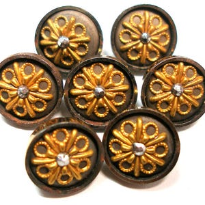 Puede incluir: Siete botones antiguos de latón con un diseño floral dorado y un centro plateado. Los botones son redondos y tienen un borde marrón oscuro. Los botones están dispuestos en un patrón circular.