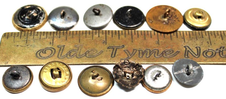 Puede incluir: Una colecci&oacute;n de botones antiguos en varios colores y dise&ntilde;os, incluyendo negro, dorado, plateado y bronce. Una regla de lat&oacute;n con el texto "Olde Tyme Not" se coloca junto a los botones. Los botones var&iacute;an en tama&ntilde;o y estilo.