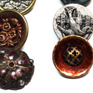 Puede incluir: Una colecci&oacute;n de botones vintage en varias formas, tama&ntilde;os y colores. Los botones presentan dise&ntilde;os intrincados, incluyendo patrones florales, un p&aacute;jaro con una valla y formas geom&eacute;tricas. La paleta de colores incluye oro, rojo, plata y negro.