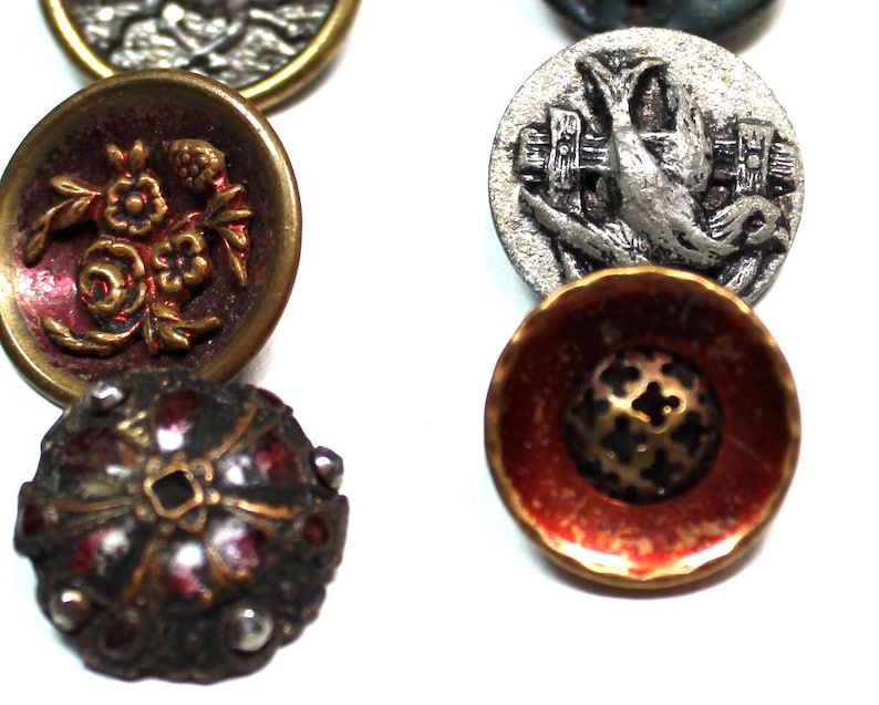 Puede incluir: Una colecci&oacute;n de botones vintage en varias formas, tama&ntilde;os y colores. Los botones presentan dise&ntilde;os intrincados, incluyendo patrones florales, un p&aacute;jaro con una valla y formas geom&eacute;tricas. La paleta de colores incluye oro, rojo, plata y negro.