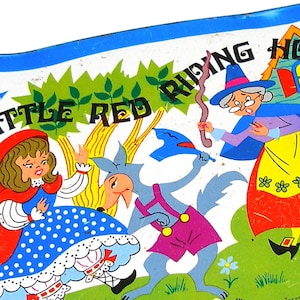 May include: A vintage tin lunchbox featuring a colourful illustration of Little Red Riding Hood, the wolf, and the grandmother. The text "Little Red Riding Hood" is printed in blue letters on a white background.