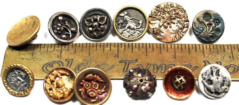 Puede incluir: Una colecci&oacute;n de botones antiguos de diversas formas, tama&ntilde;os y colores. Los botones incluyen dise&ntilde;os florales, geom&eacute;tricos y figurativos. Una regla se coloca detr&aacute;s de los botones para mostrar su tama&ntilde;o. Los botones son de metal y tienen un aspecto vintage.