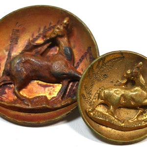 May include: Two antique metal buttons with a deer design. The larger button is a dark brown color with a deer leaping through tall grass. The smaller button is gold with a deer standing on a base.