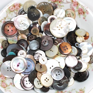 May include: A collection of various buttons in a floral patterned bowl. The buttons are in a variety of sizes, colors, and materials, including pearl, brown, black, and white. Some buttons have two holes, while others have four.