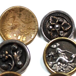 Puede incluir: Primer plano de varios botones antiguos con dise&ntilde;os detallados. Un bot&oacute;n es dorado con una escena n&aacute;utica, otro tiene un dise&ntilde;o floral plateado y un tercero presenta una figura con un tridente. Los botones var&iacute;an en tama&ntilde;o y color.