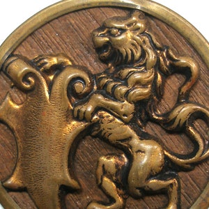 Puede incluir: Un botón de metal dorado con un diseño en relieve de un león de pie sobre sus patas traseras con la cola enroscada alrededor de su cuerpo. El león sostiene un rollo en sus patas. El botón tiene una textura de madera.