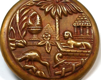 May include: A circular, antique brass button with an intricate relief design. The button depicts a palm tree, a sphinx, a pyramid, and a bird, set against a textured backdrop. The button has a slightly raised edge and a warm, golden colour.