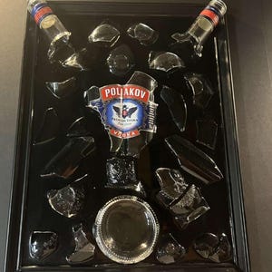 Peut inclure: Une œuvre d'art encadrée en noir représentant une bouteille de vodka Poliakov brisée. La bouteille est brisée en plusieurs morceaux, avec l'étiquette toujours visible. Les morceaux sont disposés de manière créative et artistique.