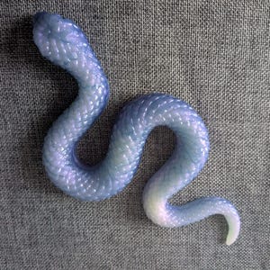 May include: A snake figurine with a translucent, iridescent finish that shifts between light blue and soft purple tones. The surface features a detailed, raised scale texture across its entire body, which is posed in a winding, S-shaped curve.