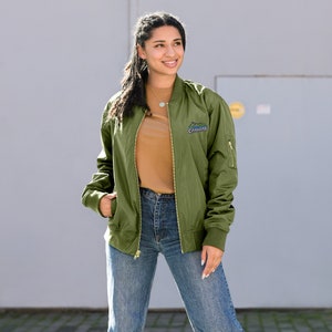 May include: Olive green bomber jacket with gold zippers and a small mountain logo that says "CANVAS". The jacket is worn over a brown t-shirt and blue jeans. The jacket has a ribbed collar, cuffs, and hem.