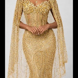 Könnte beinhalten: Ein goldenes, perlenbesetztes Abendkleid mit passendem, durchsichtigem Umhang. Das Kleid hat ein eng anliegendes Oberteil mit floralen Verzierungen und eine Meerjungfrau-Silhouette. Der Umhang drapiert über den Schultern und erzeugt einen eleganten Look.