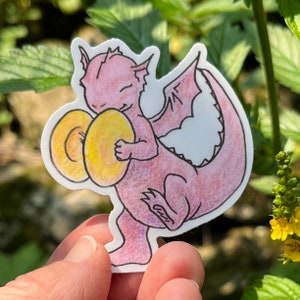 May include: A sticker of a pink dragon playing yellow cymbals. The dragon has white wings and a black outline. The sticker is held in a hand against a blurred green background.