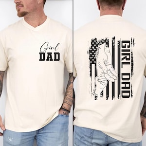 May include: Cream-colored t-shirt with the words "Girl Dad" printed on the front. The back features a black and white graphic of a hand holding a smaller hand, with the words "Girl Dad" and names printed vertically.