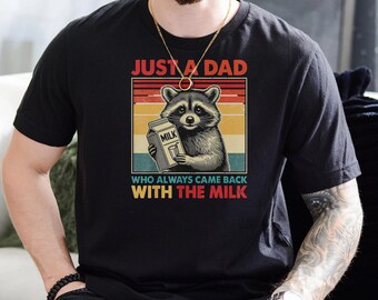 Pode incluir: Uma camiseta preta com um gráfico de estilo vintage de um guaxinim segurando uma caixa de leite. O texto "Just a Dad who always came back with the milk" está impresso abaixo do gráfico.