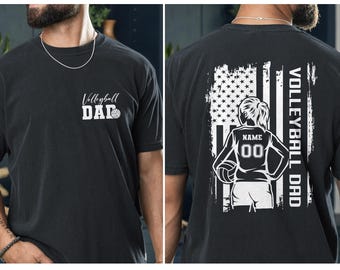 May include: Black t-shirt with "Volleyball Dad" printed on the front and a volleyball graphic. The back features an American flag design with a volleyball player graphic, the text "Volleyball Dad", and space for a name and number.