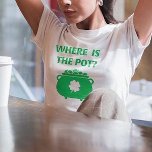 Puede incluir: Una camiseta blanca con el texto "Where is the pot?" y una olla de oro verde con un trébol.