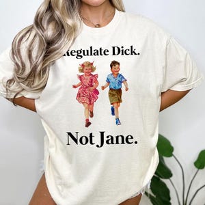 Può includere: Una maglietta bianca con una grafica di due bambini che corrono. Il testo "Regulate Dick. Not Jane." è stampato sulla maglietta.
