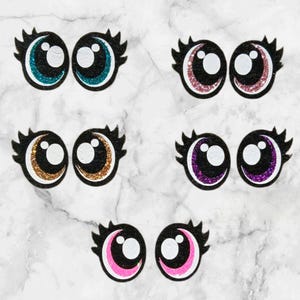 May include: Six pairs of googly eyes with different coloured glitter accents. The eyes are black with white pupils and eyelashes. The glitter colours are teal, pink, gold, purple, and pink.