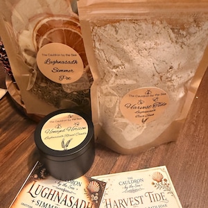 May include: Two tan pouches containing dried botanicals and bath soak, a small black candle tin, and two informational cards. The labels read The Cauldron by the Sea, Lughnasadh Simmer Pot, Harvest Tide Bath Soak, and Honeyed Horizon Ritual Candle.