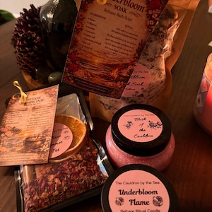 May include: An assortment of self-care products, including bath soaks, ritual candles, and a simmer pot. The items are packaged in clear and brown bags and jars, with pink labels and floral designs. The text on the labels includes "Emberbloom Soak" and "Underbloom Flame."
