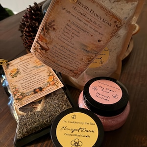 May include: An assortment of products from "The Cauldron by the Sea" on a wooden surface. Includes a bag of "Honeyed Dawn Soak" with ingredients listed, a jar of "Hey Sugar Scrub Mermaid's Shortcake", and a candle labeled "Honeyed Dawn Ostara Ritual Candle".