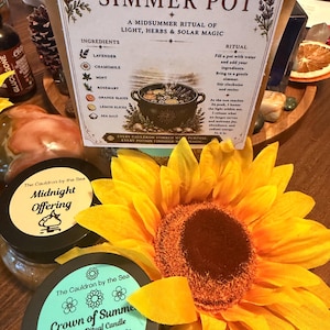 May include: A Litha Ritual Simmer Pot pouch with ingredients listed, a Midnight Offering jar, a Crown of Summer Litha Ritual Candle in a black tin, and a large yellow sunflower. The pouch text includes ingredients like lavender, chamomile, mint, rosemary, orange, lemon, and sea salt.