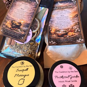 May include: An assortment of ritual products, including a simmer pot, bath soak, and candles. The simmer pot and bath soak come in decorative packaging with ingredient lists and rituals. The candles are in black tins with yellow and pink labels, featuring the names "Sunspell Meringue" and "Frostkissed Garden."