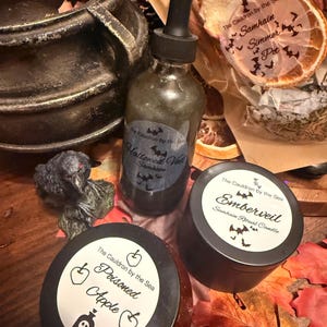Könnte beinhalten: Eine Sammlung von Gegenständen, darunter ein schwarzer Kessel, eine Glastropfflasche mit der Aufschrift Hallowed Veil Samhain, eine schwarze Dose mit der Aufschrift Emberveil Samhain Ritual Candle und ein Glas mit der Aufschrift Poisoned Apple.