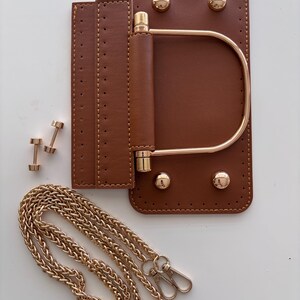 Puede incluir: Componentes de bolso de cuero marr&oacute;n con herrajes dorados. Incluye una base rectangular con esquinas redondeadas, un asa y una correa de cadena. Tambi&eacute;n se muestran dos peque&ntilde;os remaches dorados y una solapa con costuras.