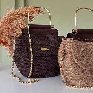 Puede incluir: Dos bolsos artesanales. Uno es marr&oacute;n oscuro, el otro es beige. Ambos tienen una textura tejida, correas de cadena doradas y asas doradas. Los bolsos tienen un cierre de solapa con un peque&ntilde;o acento dorado. El fondo es verde claro.