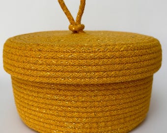 Puede incluir: Una pequeña cesta de almacenamiento redonda y amarilla con textura tejida y un asa de cuerda.