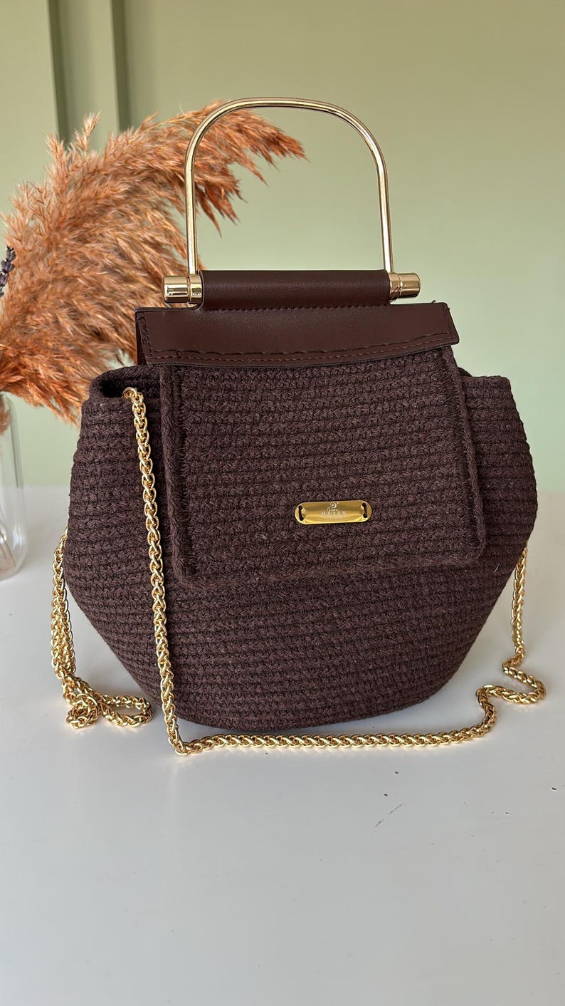 Puede incluir: Un bolso tejido marr&oacute;n con un asa y una correa de cadena doradas. El bolso tiene un cierre de solapa con un adorno dorado. La parte superior del bolso es de cuero marr&oacute;n. El bolso est&aacute; sobre una superficie blanca.