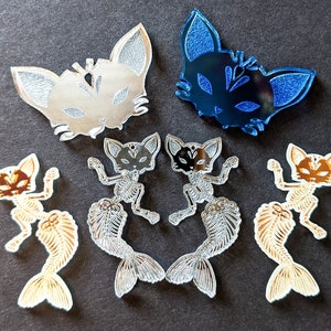 May include: Assortment of five cat-shaped acrylic charms. Two charms are silver with a cat face design, one is blue with a cat face design, and two are gold with a cat skeleton design.