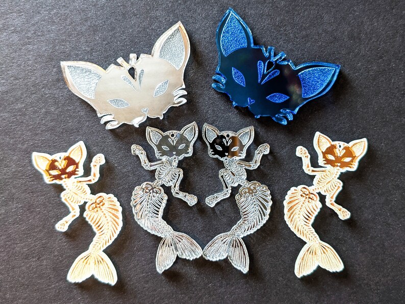 May include: Assortment of five cat-shaped acrylic charms. Two charms are silver with a cat face design, one is blue with a cat face design, and two are gold with a cat skeleton design.