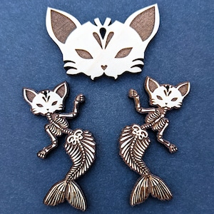 May include: Two wooden cat mermaid ornaments with a white wooden cat mask. The mermaids are skeletal and have a fish tail. The cat mask has a white face with brown ears and a black design.