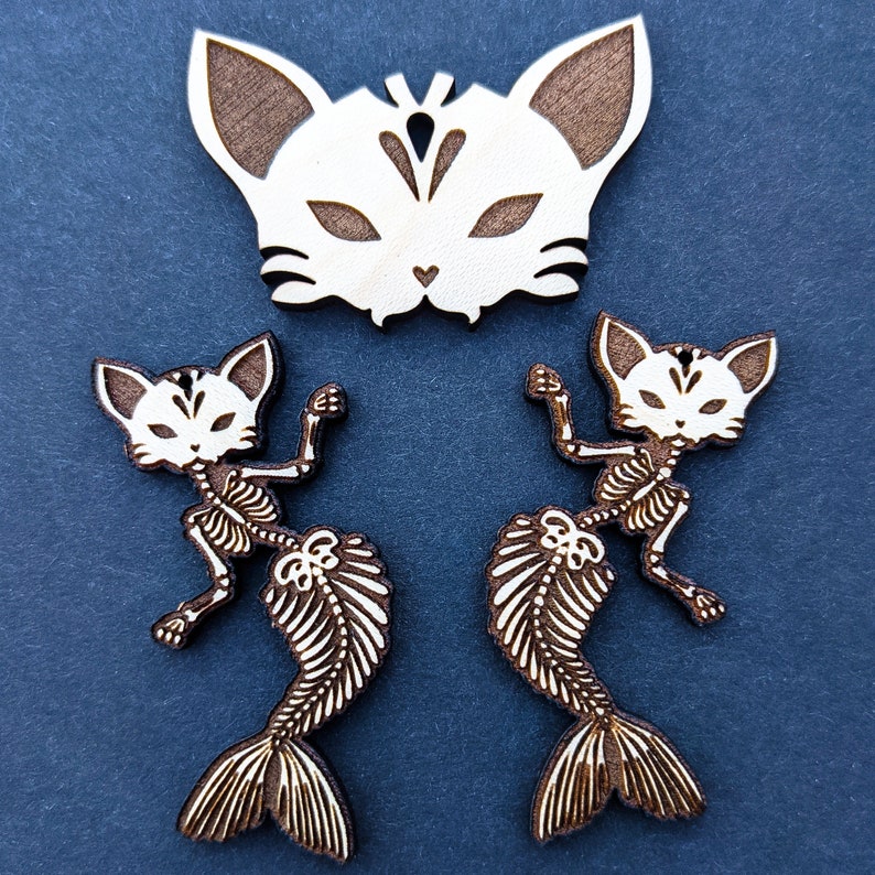 May include: Two wooden cat mermaid ornaments with a white wooden cat mask. The mermaids are skeletal and have a fish tail. The cat mask has a white face with brown ears and a black design.